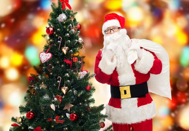 Kerstmis, vakantie en mensen concept - man in kostuum van de kerstman met tas en kerstboom stil gebaar maken over rode lichten achtergrond