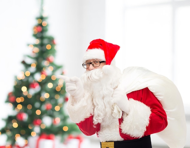Kerstmis, vakantie en mensen concept - man in kostuum van de kerstman met zak wijzende vinger over woonkamer met boom