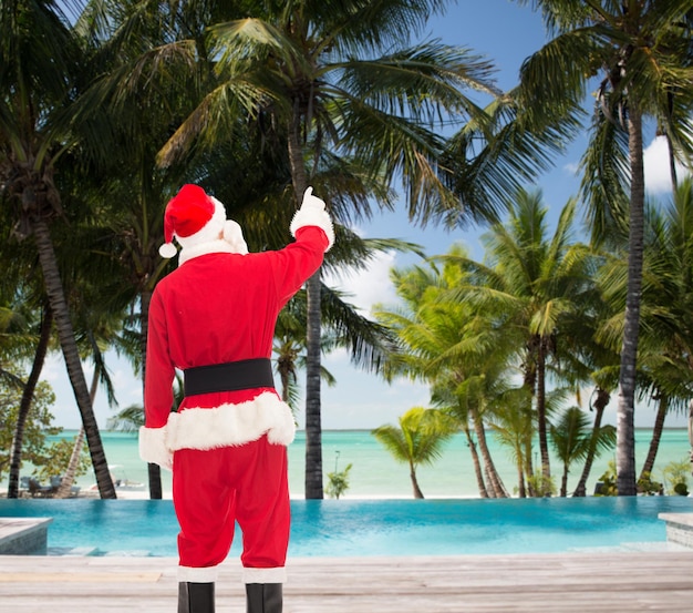 Kerstmis, vakantie en mensen concept - man in kostuum van de kerstman wijzende vinger van achteren over tropisch strand en zwembad achtergrond