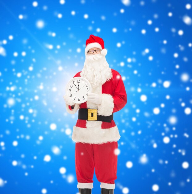 Kerstmis, vakantie en mensenconcept - man in kostuum van de kerstman met klok die twaalf toont over blauwe besneeuwde achtergrond