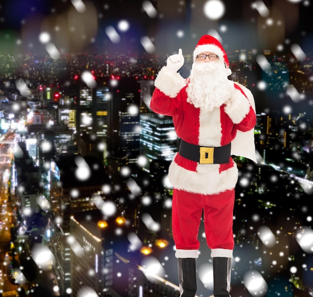 Kerstmis, vakantie, gebaar en mensen concept - man in kostuum van de kerstman met zak wijzende vinger omhoog over besneeuwde nacht stad achtergrond