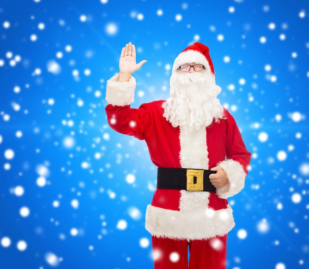 Kerstmis, vakantie, gebaar en mensenconcept - man in kostuum van de kerstman die hand over blauwe besneeuwde achtergrond zwaait