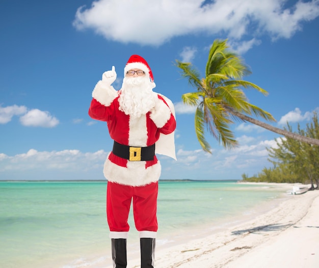 Kerstmis, vakantie, gebaar, reizen en mensen concept - man in kostuum van de kerstman met tas wijzende vinger omhoog over tropisch strand achtergrond