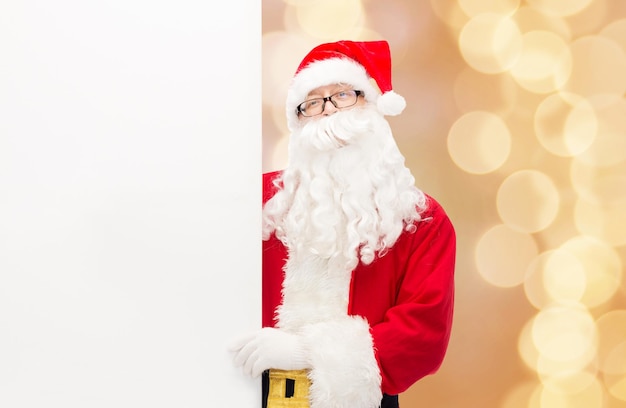 Kerstmis, vakantie, reclame en mensenconcept - man in kostuum van de kerstman met wit leeg reclamebord over beige lichtenachtergrond