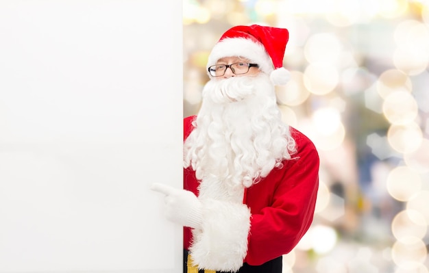 Kerstmis, vakantie, reclame, gebaar en mensenconcept - man in kostuum van de kerstman wijzende vinger naar wit leeg reclamebord over lichtenachtergrond