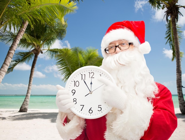 Kerstmis, vakantie, reizen en mensenconcept - man in kostuum van de kerstman met klok die twaalf toont over tropisch strandachtergrond