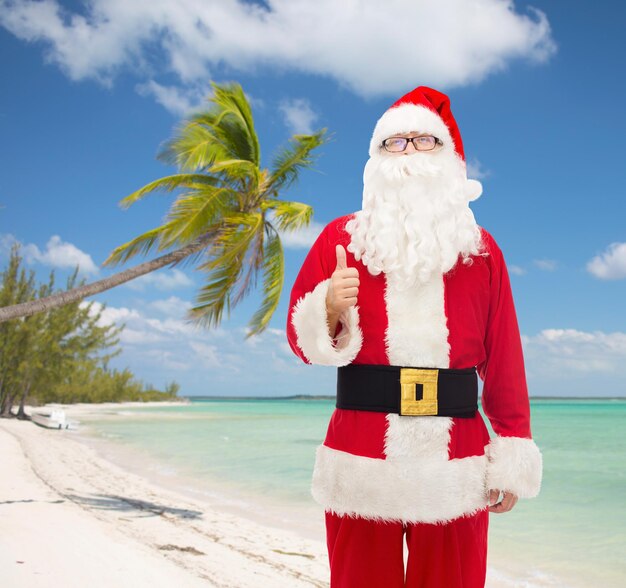 Kerstmis, vakantie, reizen, gebaar en mensen concept-man in kostuum van de kerstman duimen opdagen over tropisch strand achtergrond