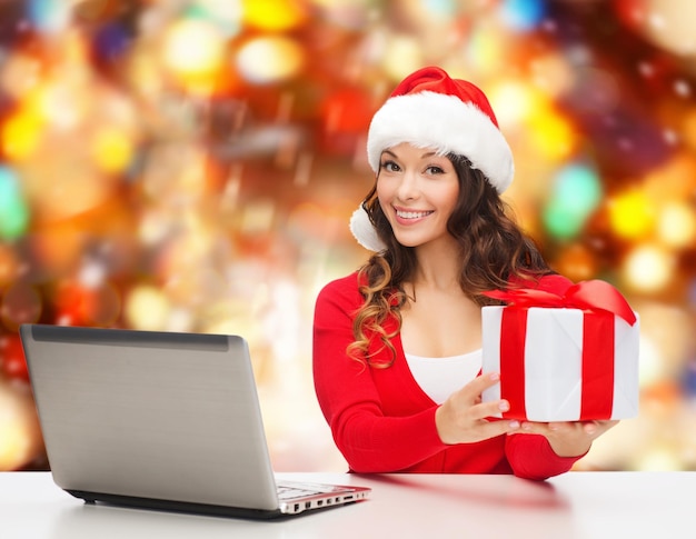 Kerstmis, vakantie, technologie en mensen concept - glimlachende vrouw in santa helper hoed met geschenkdoos en laptopcomputer over rode sneeuwt achtergrond