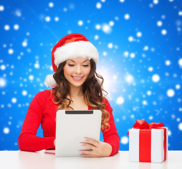 Kerstmis, vakantie, technologie en mensen concept - glimlachende vrouw in santa helper hoed met geschenkdoos en tablet pc-computer over blauwe besneeuwde achtergrond