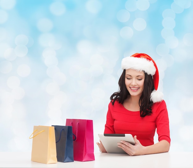 Kerstmis, vakantie, technologie en mensenconcept - glimlachende vrouw in de hoed van de Kerstmanhelper met het winkelen zakken en de computer van tabletpc over blauwe lichtenachtergrond
