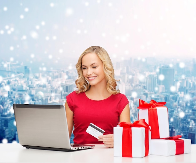 Kerstmis, vakantie, technologie en winkelconcept - glimlachende vrouw in rood leeg overhemd met giftdozen, creditcard en laptop computer over blauwe lichtenachtergrond