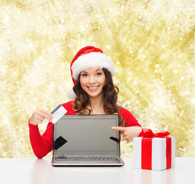 Kerstmis, vakantie, technologie en winkelen concept - lachende vrouw in santa helper hoed met geschenkdoos, creditcard en laptop computer wijzende vinger over gele lichten achtergrond