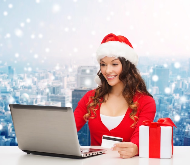 Kerstmis, vakantie, technologie en winkelen concept - lachende vrouw in santa helper hoed met geschenkdoos, creditcard en laptopcomputer over besneeuwde stad achtergrond