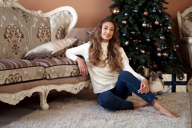 Kerstmis. Vrouw gekleed in witte trui en spijkerbroek zittend op de vloer in de buurt van kerstboom met huidige doos