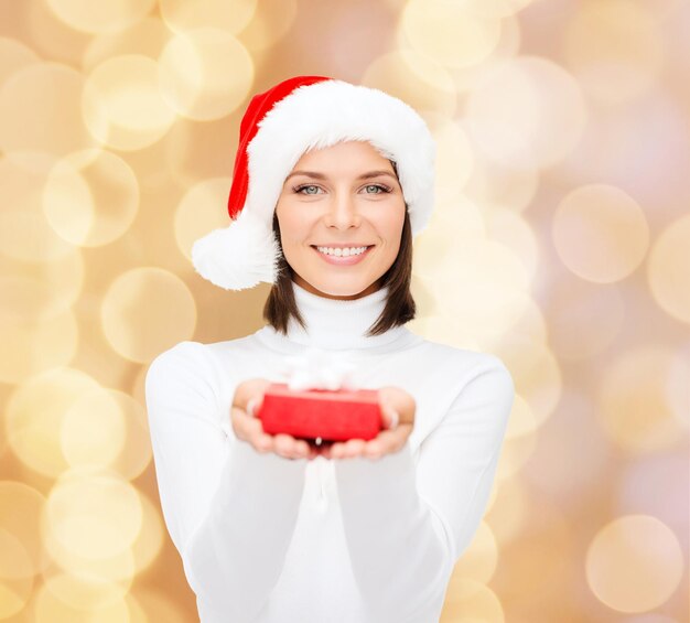 Kerstmis, winter, geluk, vakantie en mensenconcept - glimlachende vrouw in santahelperhoed met kleine rode giftdoos over beige lichtenachtergrond