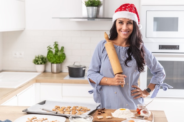 Kerstmoeder draagt een kerstmuts en houdt de roller vrolijk vast terwijl ze kerstkoekjes bakt in haar keuken.