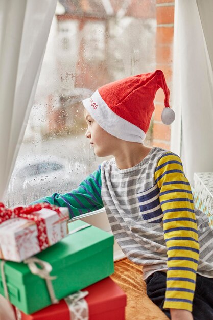 Kerstochtend in een gezinswoning Een jongen met een kerstmuts kijkt uit een slaapkamerraam