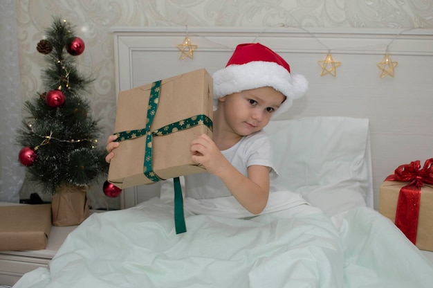 Kerstochtend, klein meisje in pyjama met geschenken in bed tegen de achtergrond van de kerstboom. Een gelukkig, glimlachend kind opent thuis het nieuwjaarscadeau. Het concept van vakantie, Nieuwjaar, Kerstmis