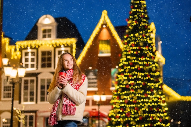 Kerstportret van een mooi meisje in een warme sjaal die op straat staat, versierd voor het nieuwe jaar en haar handen opwarmt met een deja-papieren beker met warme koffie