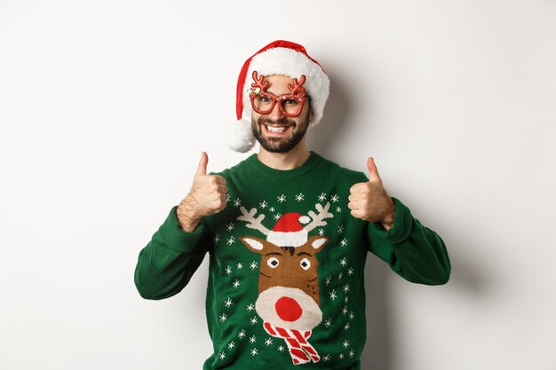 Kerstvakantie, viering concept. Gelukkige man in kerstmuts en feestbril die duimen omhoog laat zien in goedkeuring, zoals iets, staande op een witte achtergrond