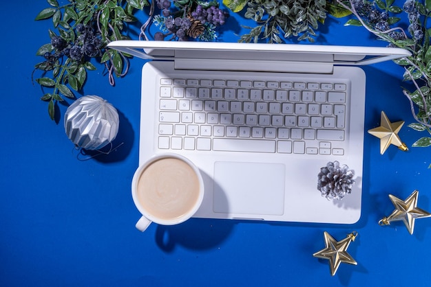 Kerstverkoop voorbereiding voor de feestdagen concept Winter kantoor werkplaats achtergrond Laptop warme chocolade latte beker met kerst speelgoed winter boom takken plat liggen op helderblauwe achtergrond