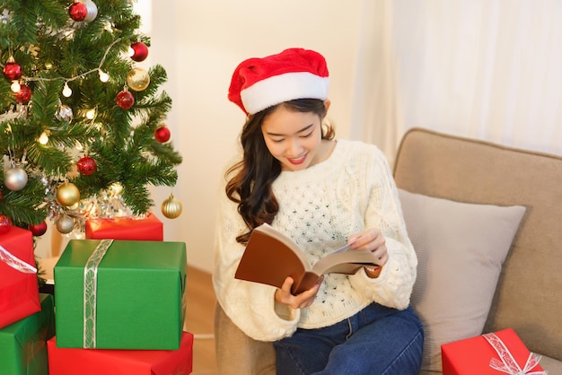 Kerstviering concept Jonge Aziatische vrouw zit op de bank om een boek te lezen in de buurt van de kerstboom