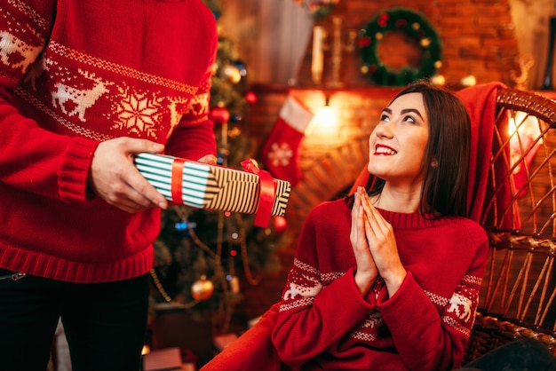 Kerstviering, mannelijke persoon maakt geschenk aan mooie jonge vrouw, dennenboom met decoratie. Gelukkig liefdepaar viert kerstvakantie