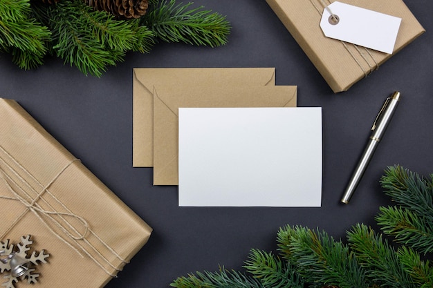 Kerstwenskaart en envelopmodel met handgemaakte geschenkdoos, dennenappels en dennenboomtakken op donkere achtergrond. feestelijke gestileerde compositie. bovenaanzicht.