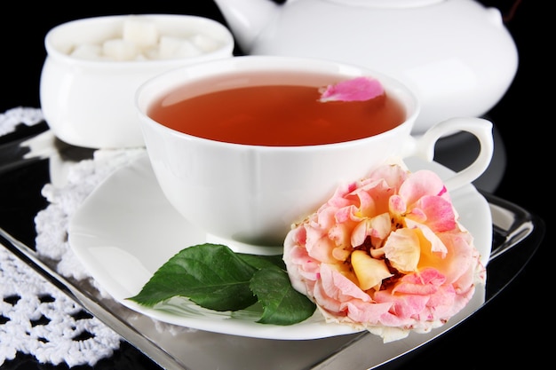 Kettle and cup of tea from tea rose on metallic tray on napkin black background