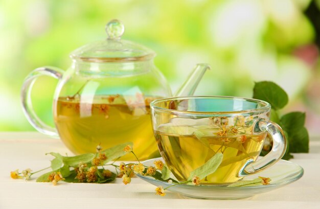 Kettle and cup of tea with linden on wooden table on nature background