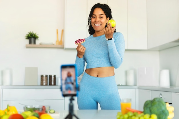 Keuze uit gezond of junkfood Fit zwarte vrouw die video opneemt op mobiele telefoon met donut en appelkeukeninterieur