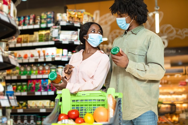 Keuze van de consument. Afrikaans familiepaar dat voedsel kiest om boodschappen te doen in de supermarkt, potten vast te houden en met de vingers te wijzen, beschermende gezichtsmaskers te dragen. Echtgenoten die eten kopen in de winkel