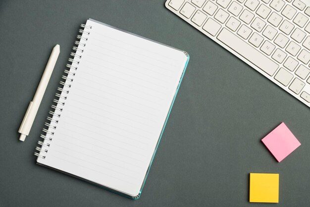 Keyboard over a table beside a notebook and pens with sticky notes computer keypad on top of a