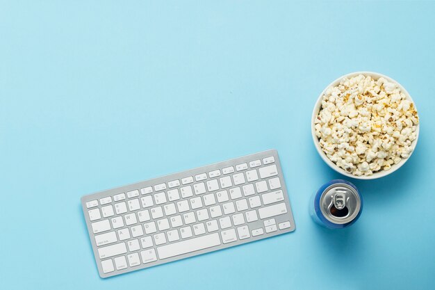 Keyboard and tin can with a drink, energy drink, a bowl of popcorn on a blue background. The concept of watching movies, TV shows, sports events online. Flat lay, top view