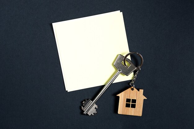 Keychain in the shape of wooden house with key on a black background with a square sheet for notes. Building, design, project, moving to new house, mortgage, rent and purchase real estate. Copy space