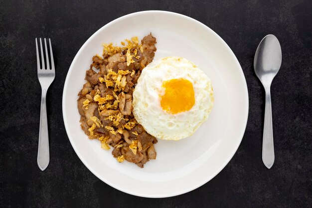 Khao Moo Tod Kratiam Kai Dao Thai food streamed rice topped with fried pork with garlic and fried egg in ceramic plate beside fork and spoon on dark tone texture background