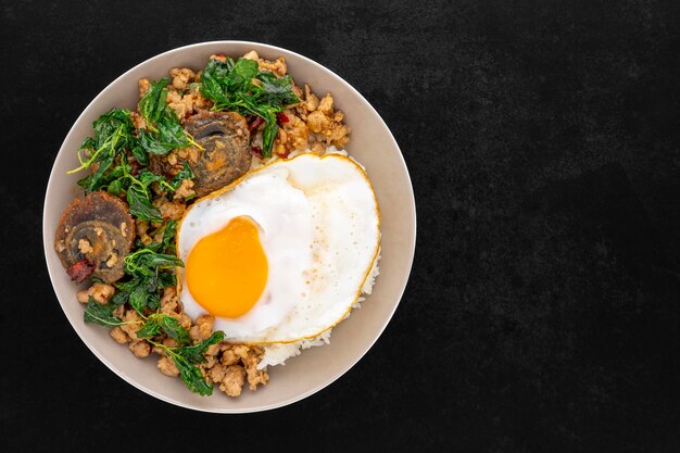 Khao Pad Ka Prao Kai Yeow Ma Kai Dao 태국 음식은 바질을 곁들인 밥을 볶고 세기의 계란 다진 돼지고기와 달걀 프라이를 세라믹 접시에 어두운 톤의 질감 배경 위에 올려 놓습니다.