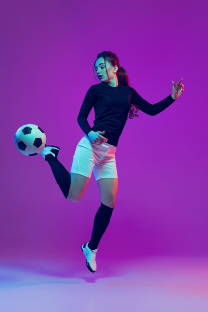 Kick ball portrait of professional female soccer player jumping isolated on purple studio background
