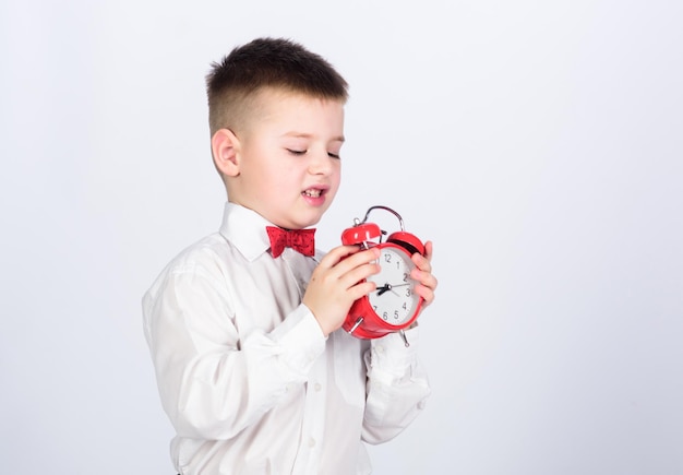 Kid adorable boy white shirt red bow tie Develop self discipline Set up alarm clock Child little boy hold red clock It is time Schedule and timing Morning routine Schoolboy with alarm clock