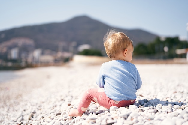 Малыш в синем свитере и красных штанах сидит, повернув голову вправо, на галечном пляже напротив