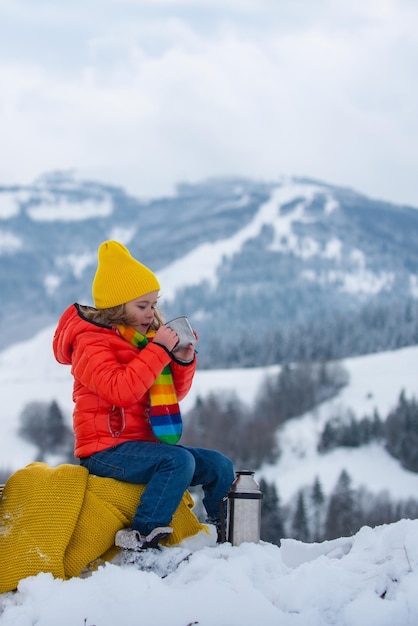 Kid boy hold mug cup with warm tea sit on sleigh ride ouside cold winter weather warm cup of hot dri