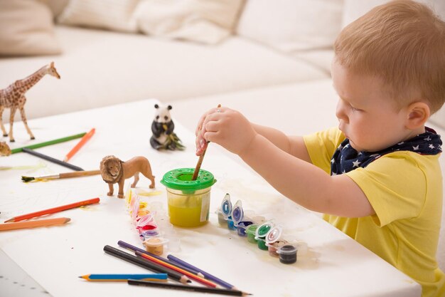 Kid Boy in geel shirt zittend aan tafel en tekenen met kleurrijke verf binnenshuis kind plezier