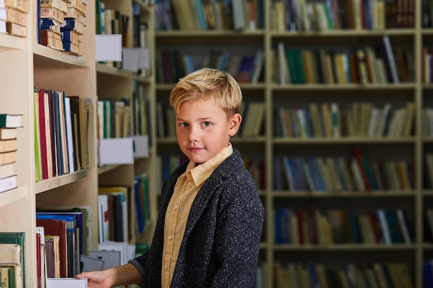 kid boy in library, looking at camera. school boy stand between shelves with books