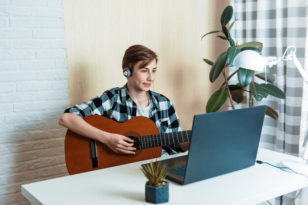 Kid boy playing guitar and watching online lessons on laptop while practicing at home. Online training, online classes