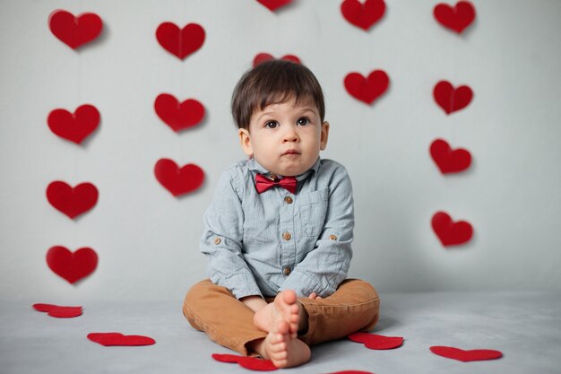 A kid boy in a shirt with a red bow tie sits against a gray wall with hearts on Valentine's Day.