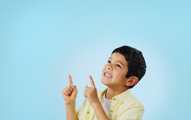Kid boy and smile while pointing in studio on blue background with mockup for announcement offer or deal Youth happy and excited about notification on social media online or digital marketing