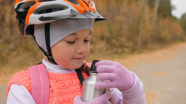 Kid drink water from an aluminum flask bottle One caucasian children rides bike road in autumn park Little girl riding black orange mtb cycle in forest Biker motion ride with backpack and helmet