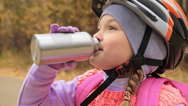 Kid drink water from an aluminum flask One caucasian children rides bike road in autumn park Little girl riding black orange mtb cycle in forest Biker motion ride with backpack and helmet