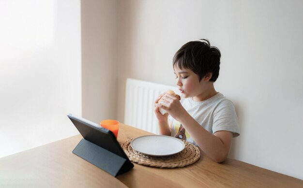 Kid eating eating homemade bacon sandwiches and watching cartoon on tablet Child boy having breakfast and playing game online on internet with friends before go to school in the morning