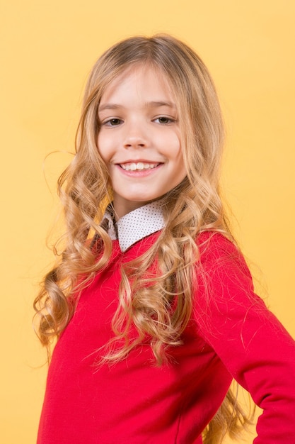 子供のファッションとスタイル。オレンジ色の背景に女の子の笑顔。美しさ、見た目、髪型。赤いセーターの巻き毛のブロンドの髪の子供。幸せな子供の頃のコンセプト。